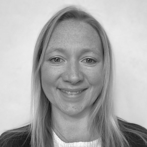 Photograph of Dee wearing a white top and black cardigan. She has long, blonde hair, light-coloured freckled skin, and a bright smile.